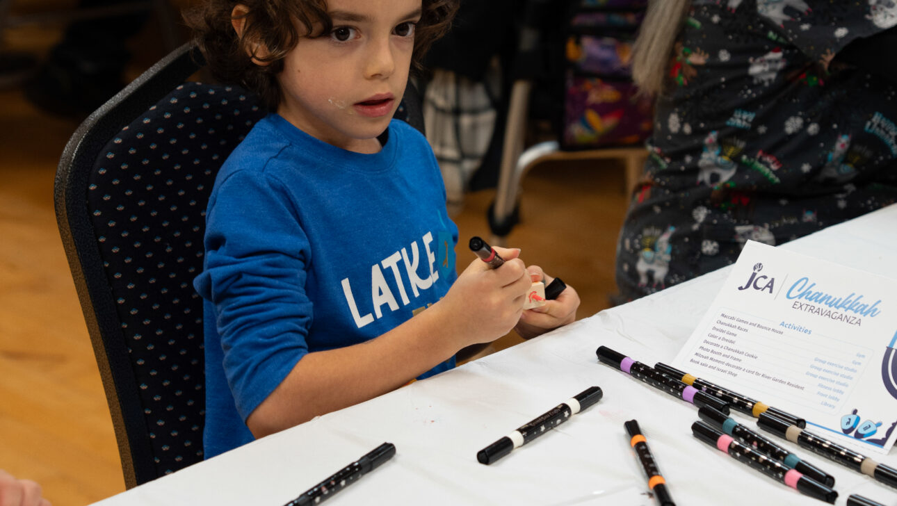 Young child wearing a blue shirt and kippah sits at a table with markers and wooden blocks, participating in an arts and crafts activity.