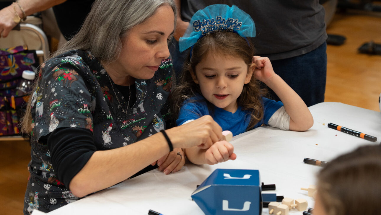 An adult helps a young girl decorate a small blue wooden house at a craft table, surrounded by markers and wooden pieces. Other people are working on crafts in the background.