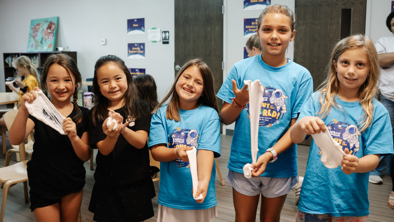 Five young girls stand indoors, smiling and holding slime. Four girls wear matching blue t-shirts, while one wears black. Classroom chairs and posters are visible in the background.
