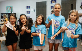 Five young girls stand indoors, smiling and holding slime. Four girls wear matching blue t-shirts, while one wears black. Classroom chairs and posters are visible in the background.