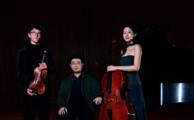Three musicians pose on stage with a violin, cello, and grand piano in a dimly lit concert hall.