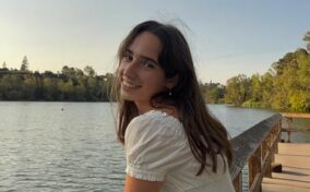 A woman in a white dress leans on a wooden railing by a calm lake, smiling at the camera with trees and a clear sky in the background.