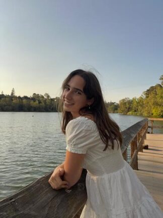 A woman in a white dress leans on a wooden railing by a calm lake, smiling at the camera with trees and a clear sky in the background.