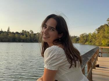 A woman in a white dress leans on a wooden railing by a calm lake, smiling at the camera with trees and a clear sky in the background.