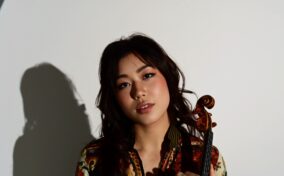 A person with long dark hair holds a violin while standing in front of a circular white background and looking at the camera.