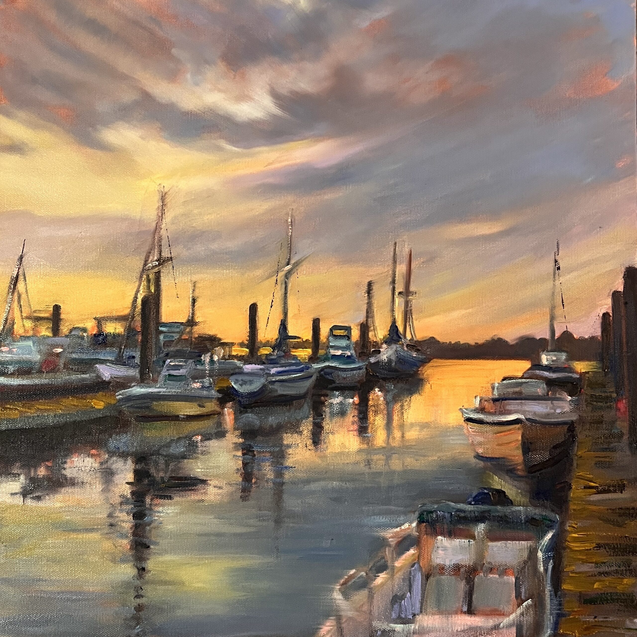 Boats are docked at a marina during sunset, with their reflections visible in the calm water and a colorful sky overhead.