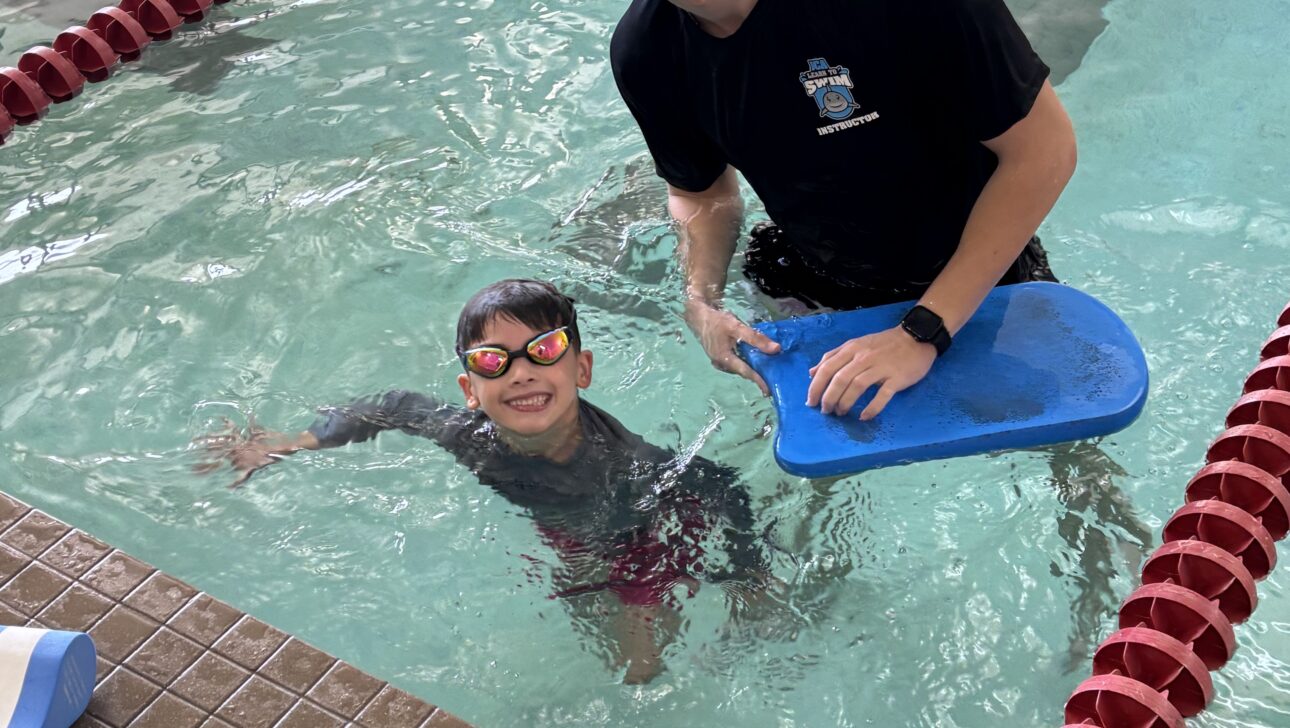 A swimming instructor holds a kickboard while smiling with a young child wearing goggles in a swimming pool near the lane lines.