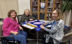 Three women sit around a game table playing mahjong. Bookshelves, a large plant, and cleaning supplies are visible in the background.