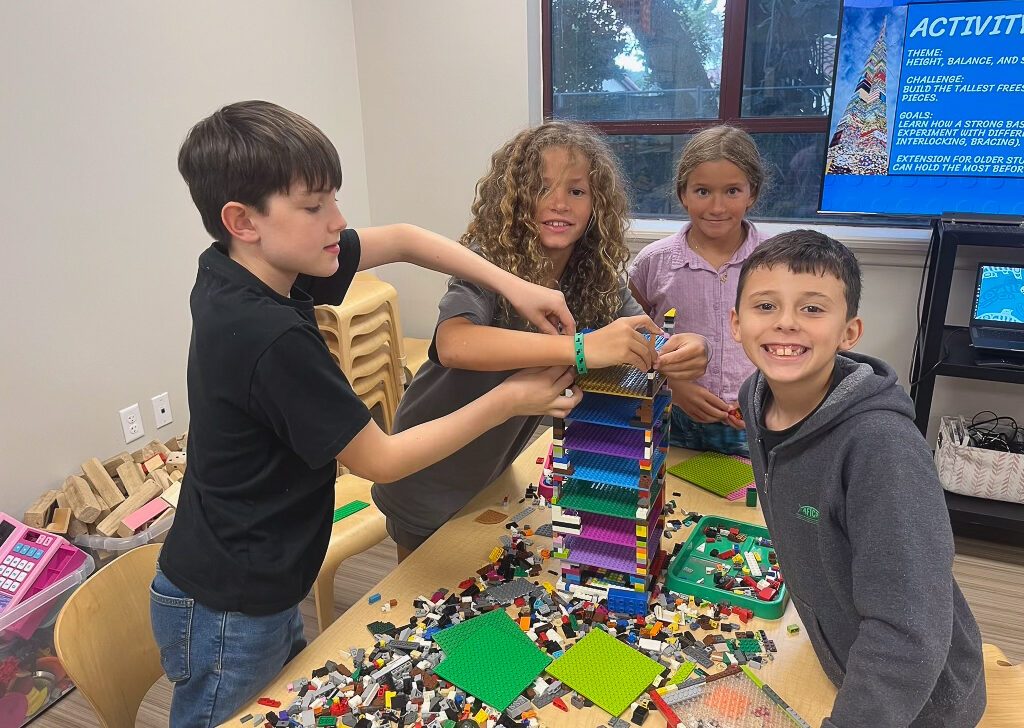 Four children build a tall structure with colorful LEGO bricks at a table, smiling and working together in a classroom setting.