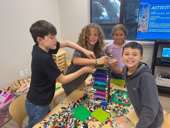 Four children build a tall structure with colorful LEGO bricks at a table, smiling and working together in a classroom setting.