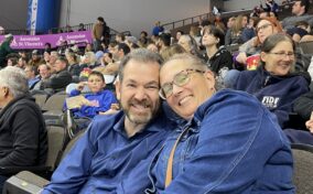 A man and a woman sit closely together and smile at a sports event, surrounded by other spectators in stadium seating.