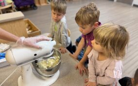 Three young children watch as an adult operates a stand mixer with butter inside on a countertop in a classroom setting.