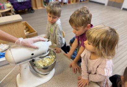 Three young children watch as an adult operates a stand mixer with butter inside on a countertop in a classroom setting.