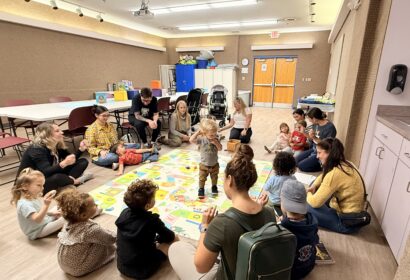 Adults and young children sit in a circle on the floor of a classroom, engaging in an activity together with toys and books visible around them.