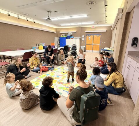 Adults and young children sit in a circle on the floor of a classroom, engaging in an activity together with toys and books visible around them.