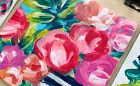 Colorful floral painting featuring pink, red, and peach flowers in a blue and white striped vase, surrounded by paint tubes and art supplies on a light wooden table.