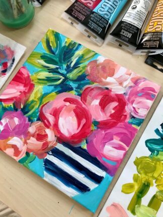 Colorful floral painting featuring pink, red, and peach flowers in a blue and white striped vase, surrounded by paint tubes and art supplies on a light wooden table.