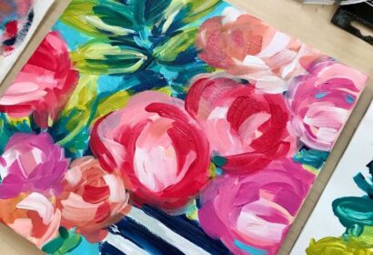 Colorful floral painting featuring pink, red, and peach flowers in a blue and white striped vase, surrounded by paint tubes and art supplies on a light wooden table.