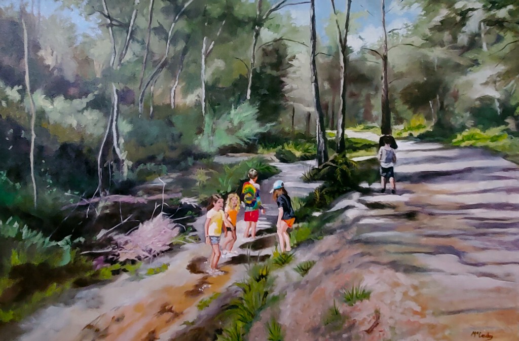 A group of children stand and wade in a shallow creek in a wooded area, while an adult walks alone on a nearby dirt path surrounded by trees and greenery.