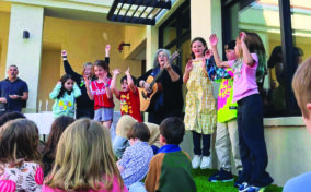 A group of children and an adult with a guitar perform outside, raising their arms, while other children sit on the grass watching.