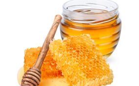 A glass jar of honey, two honeycomb pieces, and a wooden honey dipper are arranged on a white background.