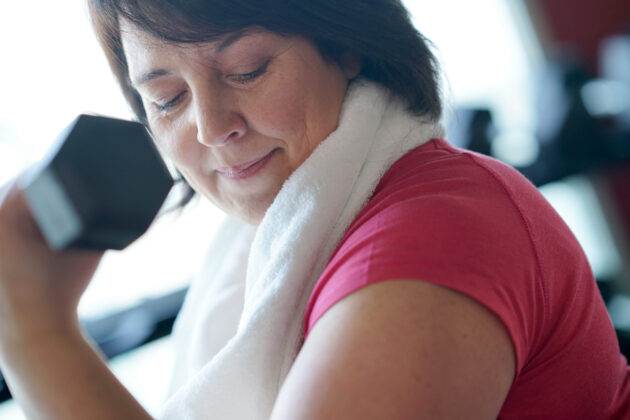 Woman in a pink shirt with a white towel over her shoulders lifts a dumbbell, looking down at her arm in a gym setting.