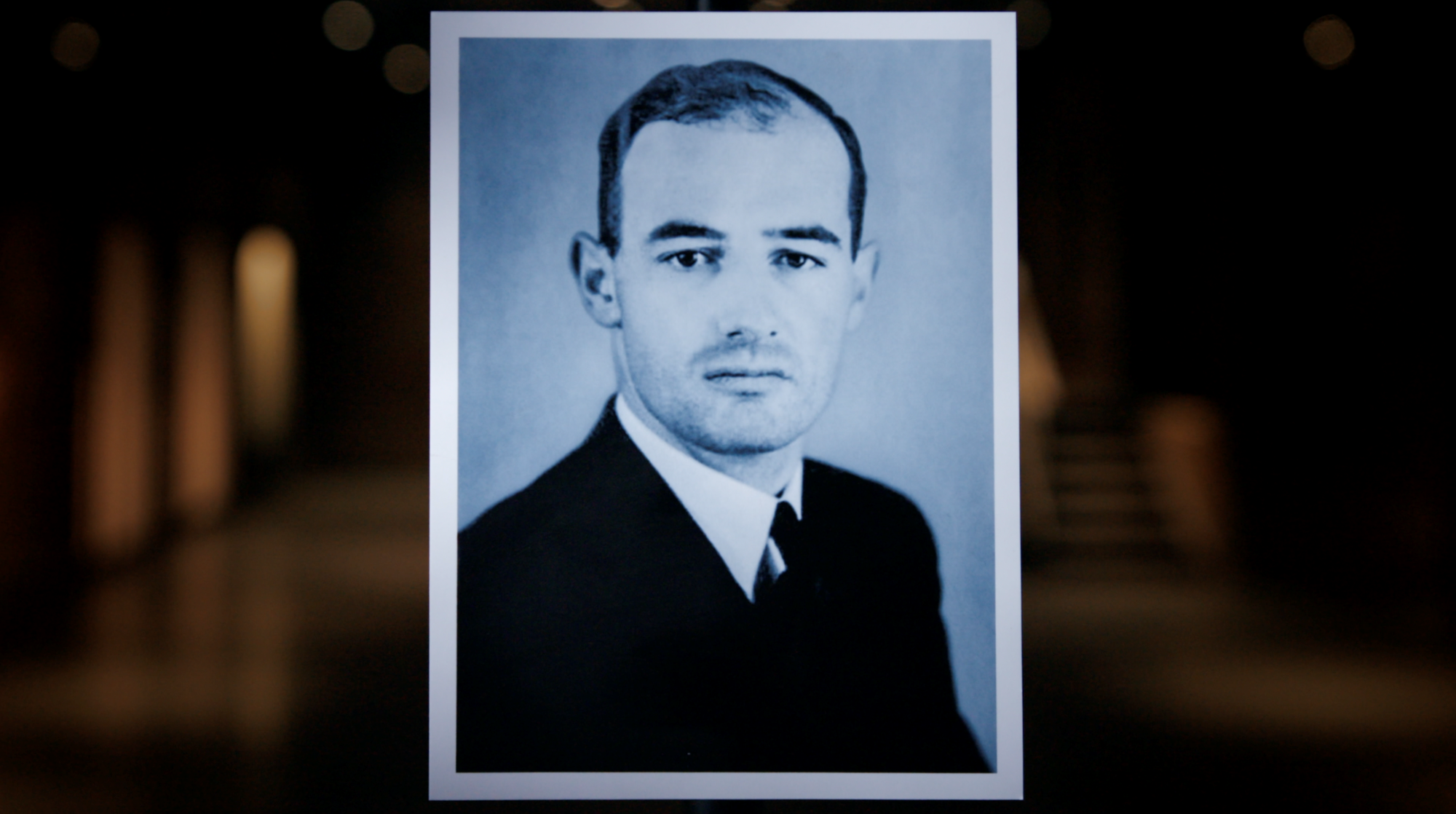 A black and white portrait photo of a man in a suit and tie, with short hair, displayed against a blurred dark background.
