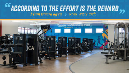 Modern gym with various exercise machines; a large sign on the wall reads, “According to the effort is the reward” in English, Hebrew, and transliteration.