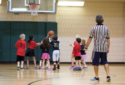 A referee watches a group of young children playing basketball in a gym.