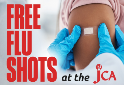 A medical professional in blue gloves places a bandage on a patient’s arm, with the text “FREE FLU SHOTS at the JCA.”